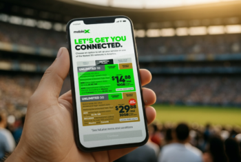 Hand holding a phone in a crowd at an outdoor sporting event. Phone screen shows MobileX new plans and prices with priority data